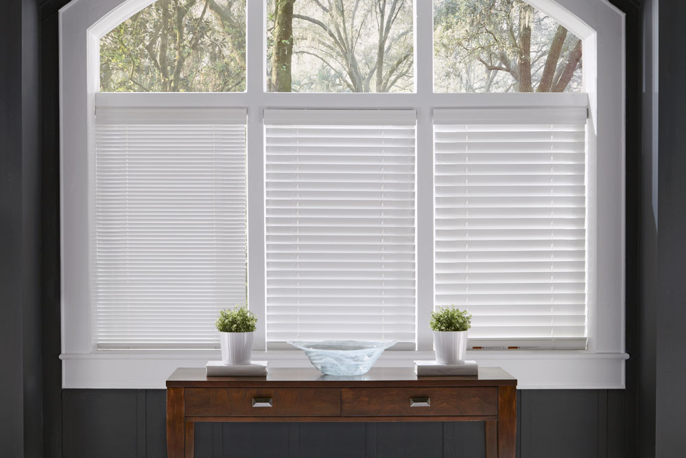 three white Heartland Woods® Wood Blinds in 1", 2" and 2 1/2" slat sizes in the open position behind a brown desk against dark gray walls