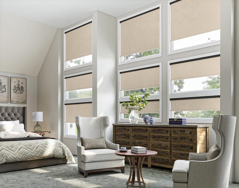 Several tan colored Genesis® Motorized Roller Shades in a bedroom with a bed and a brown dresser in front of them