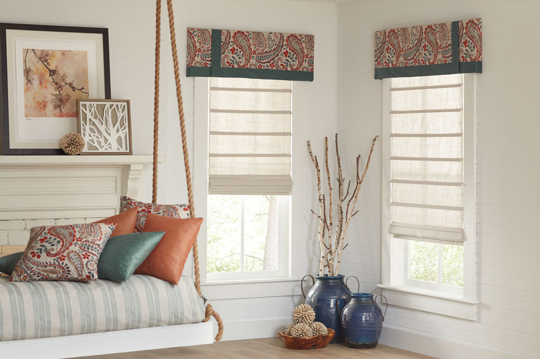 Two white Interior Masterpieces® fabric shades with floral patterned red, blue, and white fabric cornices with blue banding and custom pillows in the foreground on a swinging bench