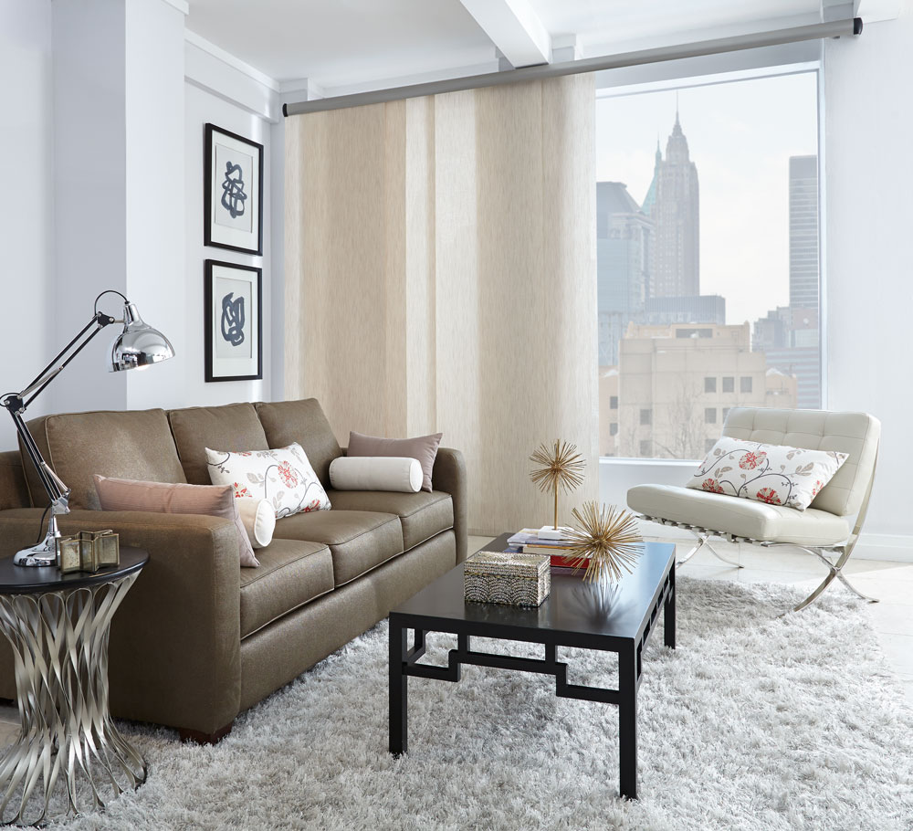 White Genesis® panel track in the open position in a picture window with a view of the city outside and a brown couch with custom Interior Masterpieces® pillows