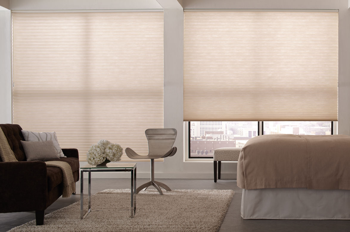 Two large picture windows in a bedroom with a dark sofa and tan bed with Parasol® Cellular Shades behind in a tan fabric