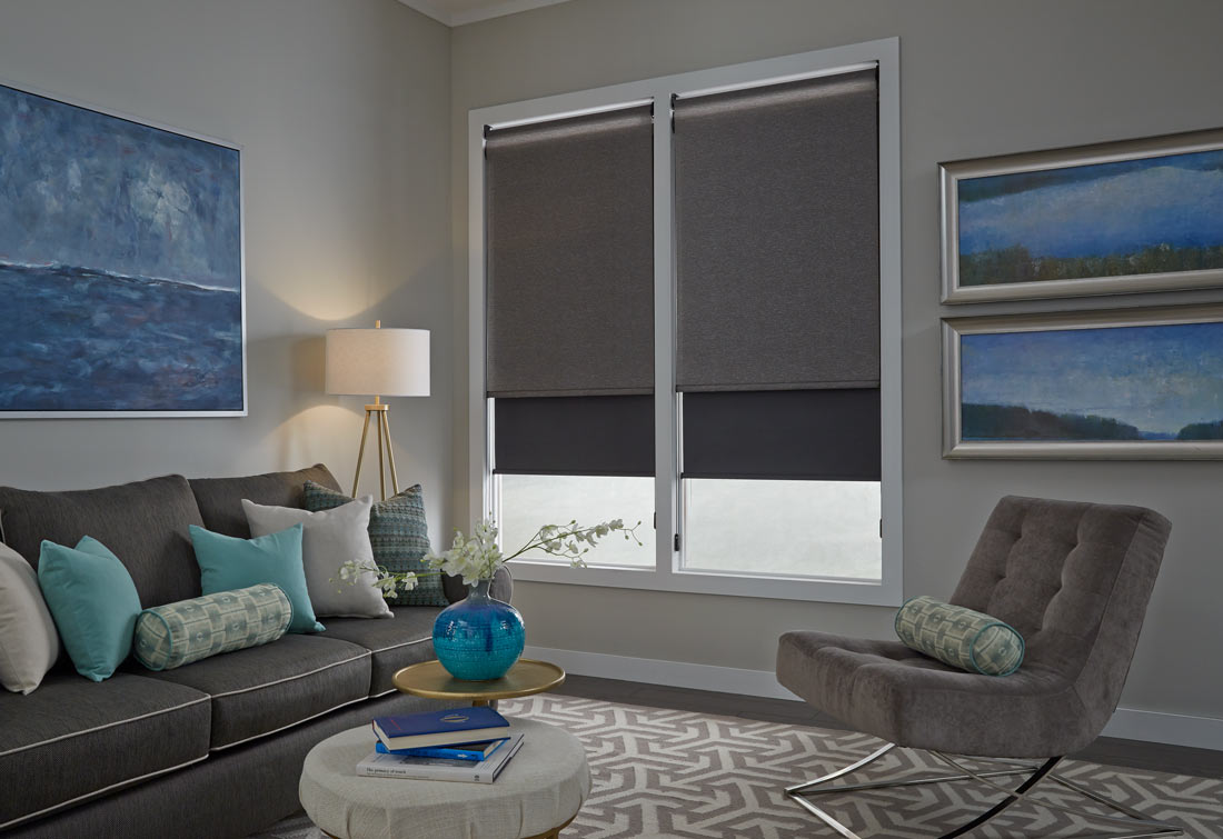 Two large gray Genesis® Dual Roller Shades behind a dark gray couch with accenting blue, gray, and green Interior Masterpieces® Custom Pillows