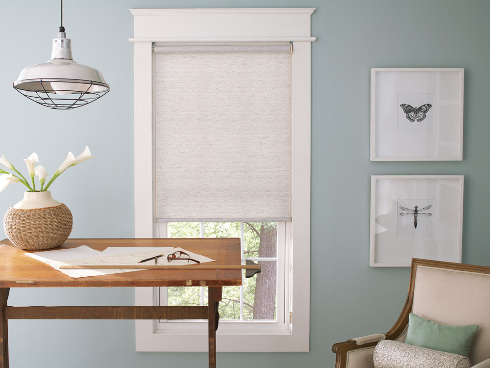 white Genesis® Custom Roller Shade in a room with light blue walls and a brown desk nearby