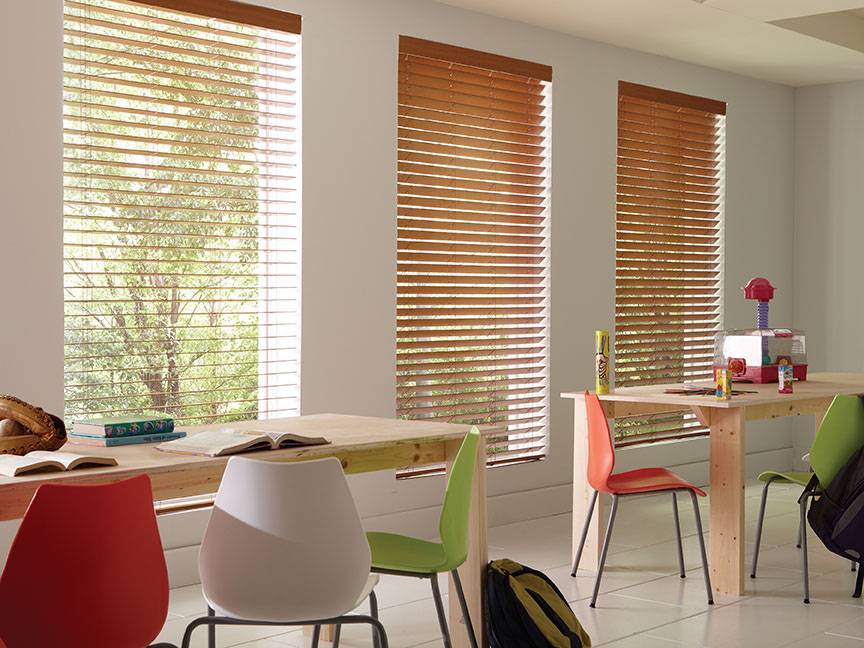 Three large custom stained faux wood blinds hang in a classroom with brightly colored chairs.