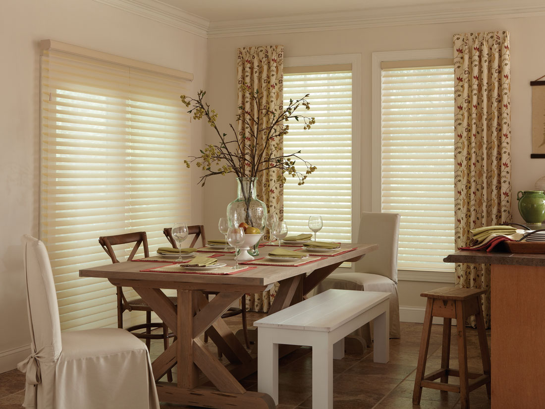 Light tan colored Tenera® sheer shades hanging over a wide patio door on one wall and two windows on the other wall with Interior Masterpieces® Draperies accenting them along with a dining room table that has Interior Masterpieces® table accessories on it