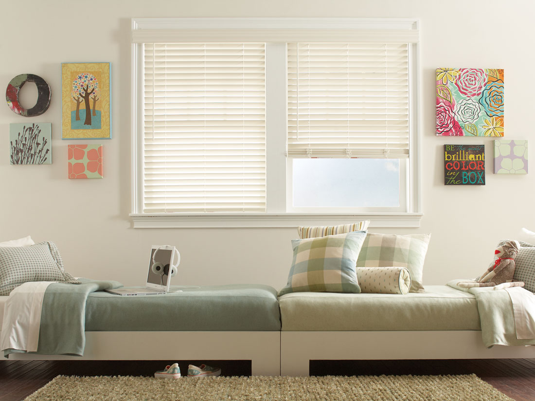 White Fidelis® Faux Wood Blinds in a bedroom with two beds in front that have light green Interior Masterpieces® Bedding and Pillows