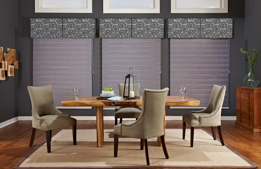 three large gray Allure® Transitional Shades with green and blue floral patterned Interior Masterpieces® fabric cornices against a dark gray wall behind a brown table and chairs