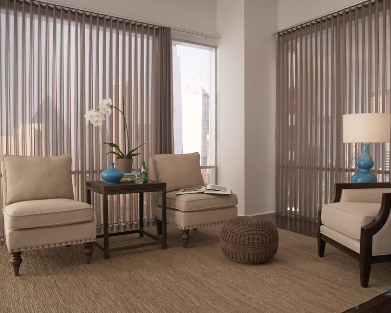 Warm gray sheer fabric wrapped vertical blinds hang in a sitting room area in a sky rise apartment with dark features, tan chairs and a brown rug.