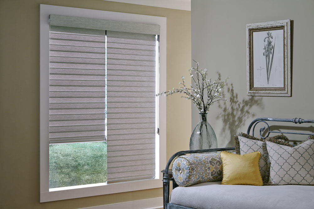 two light gray Allure® Transitional Shades under one cassette in a large window against cream colored walls witha bench that has Interior Masterpieces® Custom Pillows in yellow and a floral pattern on it
