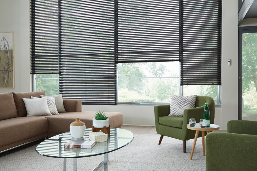 4 large windows with black 2" aluminum blinds behind a modern sofa and chair with a motorized remote hanging on the wall next to the very large blinds
