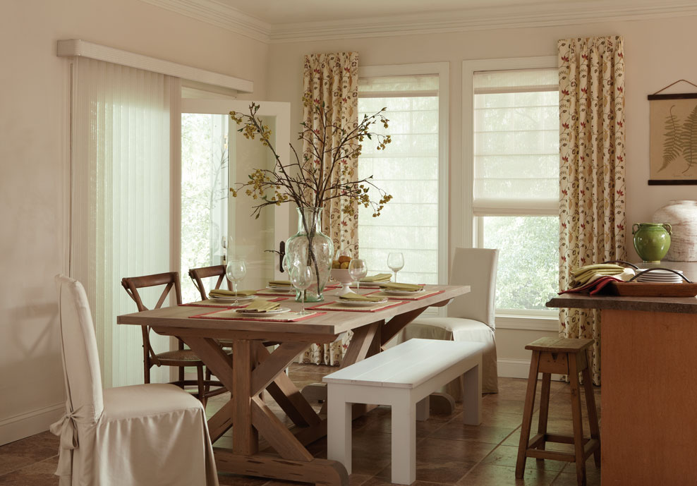 white Discoveries® Vertical Blinds on one large patio door with custom Genesis® shades with Interior Masterpieces® draperies on the other wall in a kitchen with a brown table and white covered chairs