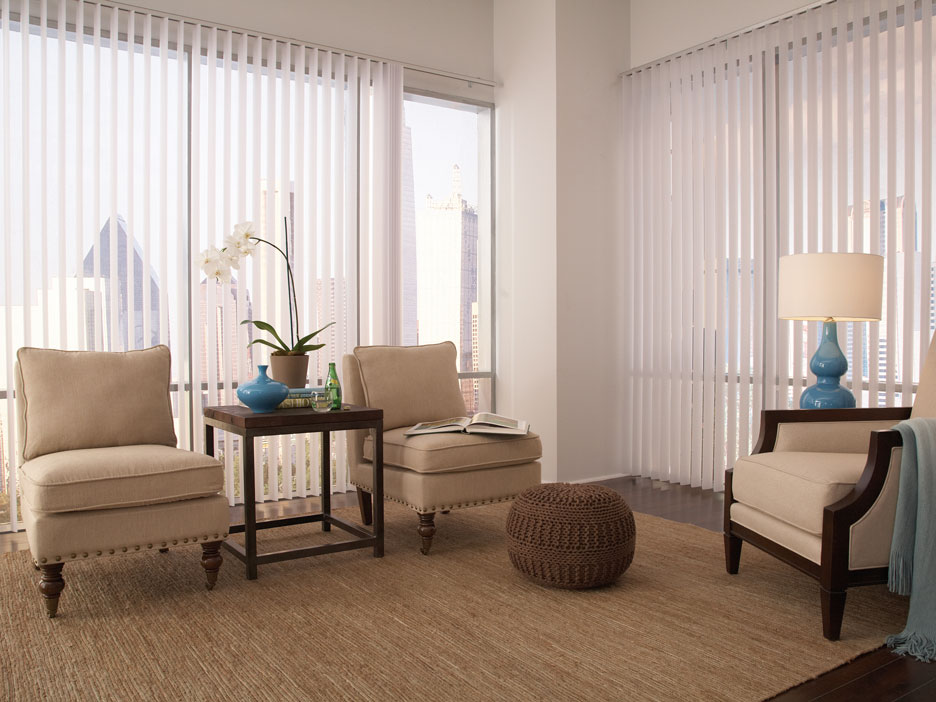 Two large picture windows with Discoveries® Vertical Blinds hanging in them in the open position in a room with tan furniture and blue accent pieces