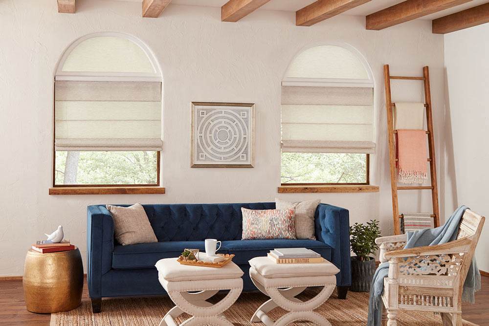 Tan colored Genesis® Hobbled Roman Shades with half circle Custom Shapes and a Palladian Shelf between behind a blue couch with tan pillows
