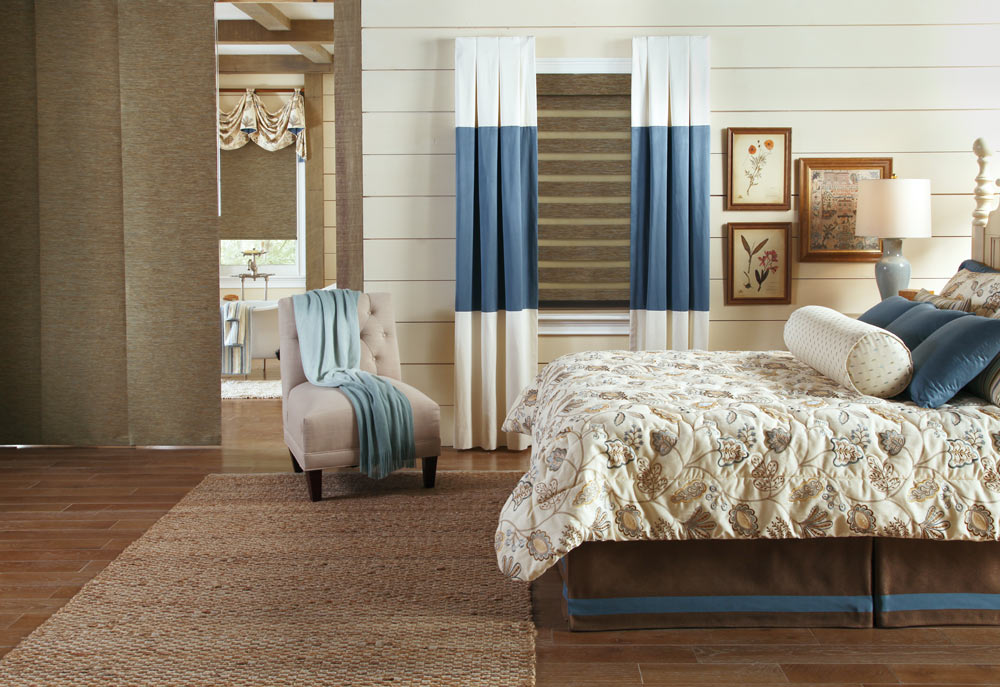 brown Allure® Transitional Shade in a window with blue and white Draperies from Interior Masterpieces® along with a Genesis® Companion Roller Shade wtih fabric valance behind a brown Panel Track with a bed that has custom bedding and pillows on it in the 