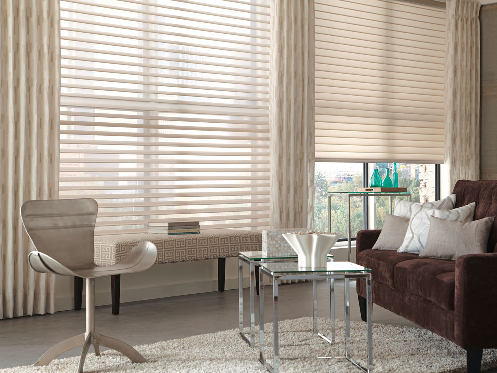 two large tan Tenera® sheer shades with custom Interior Masterpieces® draperies hanging in between them with a light background and tan geometric pattern and light custom pillows on a dark brown couch in the foreground