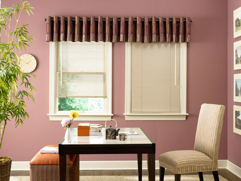 Two tan colored Classic Collection® Aluminum Blinds with a red Interior Masterpeices® Fabric Valance with custom hardware behind a black table and tan chair