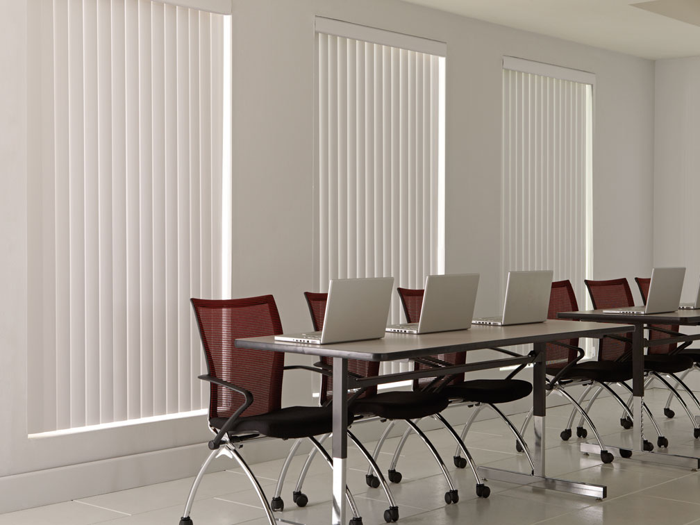 3 large windows with white colored Discoveries® Vertical Blinds in them in an office with several red chairs and laptops